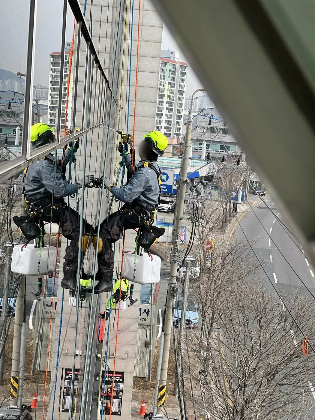 고층 건물 유리 외벽에 두 작업자가 로프로 매달려 세척하는 장면